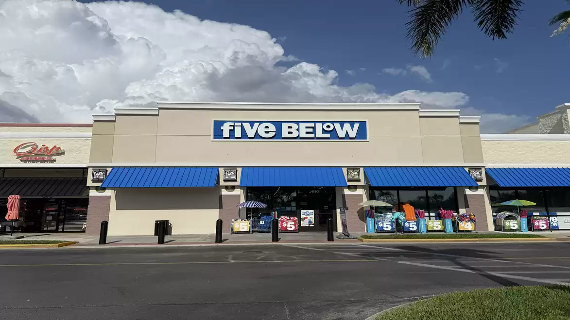 Exterior view of a Five Below store with a blue roof and signs, surrounded by tropical plants and blue sky.