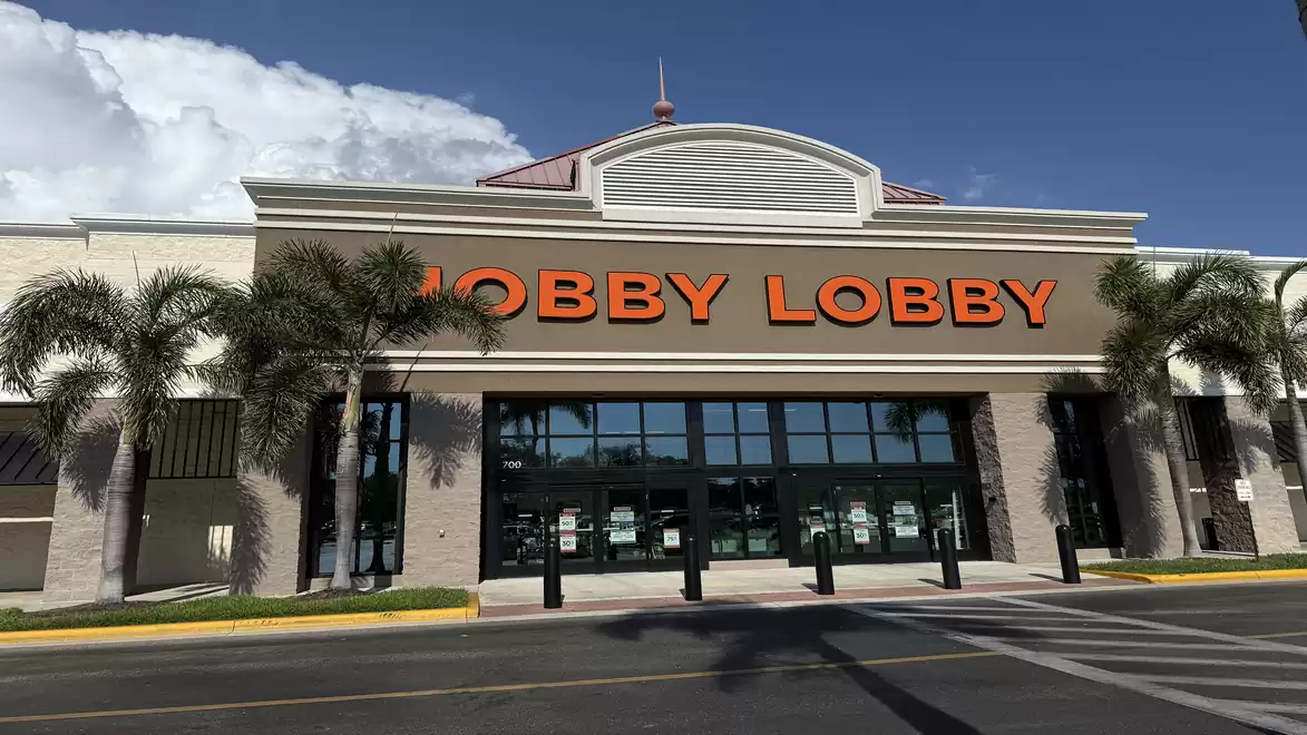 Entrance of a large retail store called Hobby Lobby with palm trees in the foreground, blue sky, and white clouds in the background.