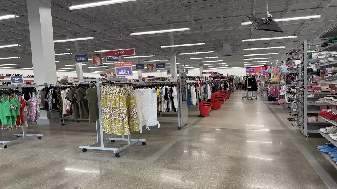 A large clothing store with numerous racks filled with various garments like shirts and dresses. In the background, red shopping baskets and more shelves with products are visible.