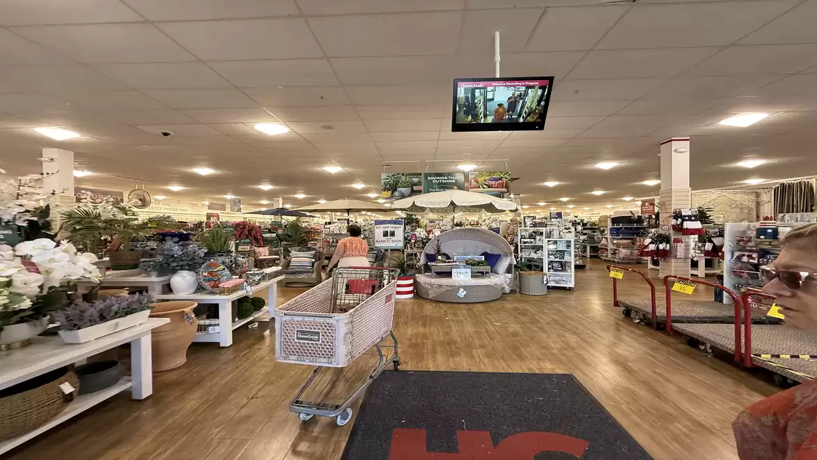 Interior view of a well-lit shopping center with shelves full of goods, shopping carts, and a large mirror screen on the ceiling.