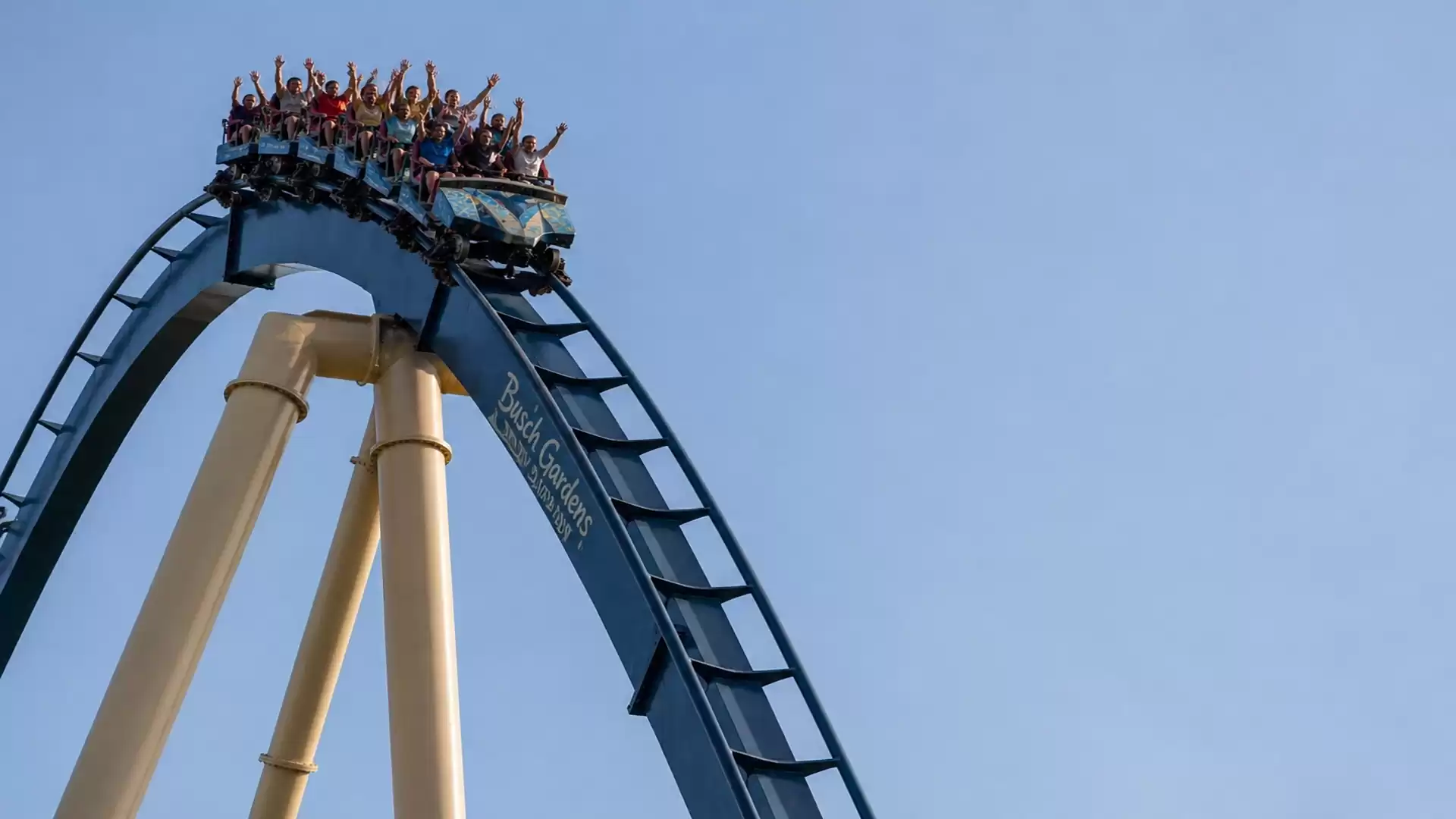 A roller coaster with several people reaches the highest point of the track before the descent with a clear blue sky in the background.