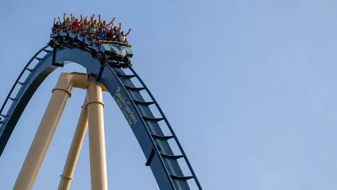 A roller coaster with several people reaches the highest point of the track before the descent with a clear blue sky in the background.