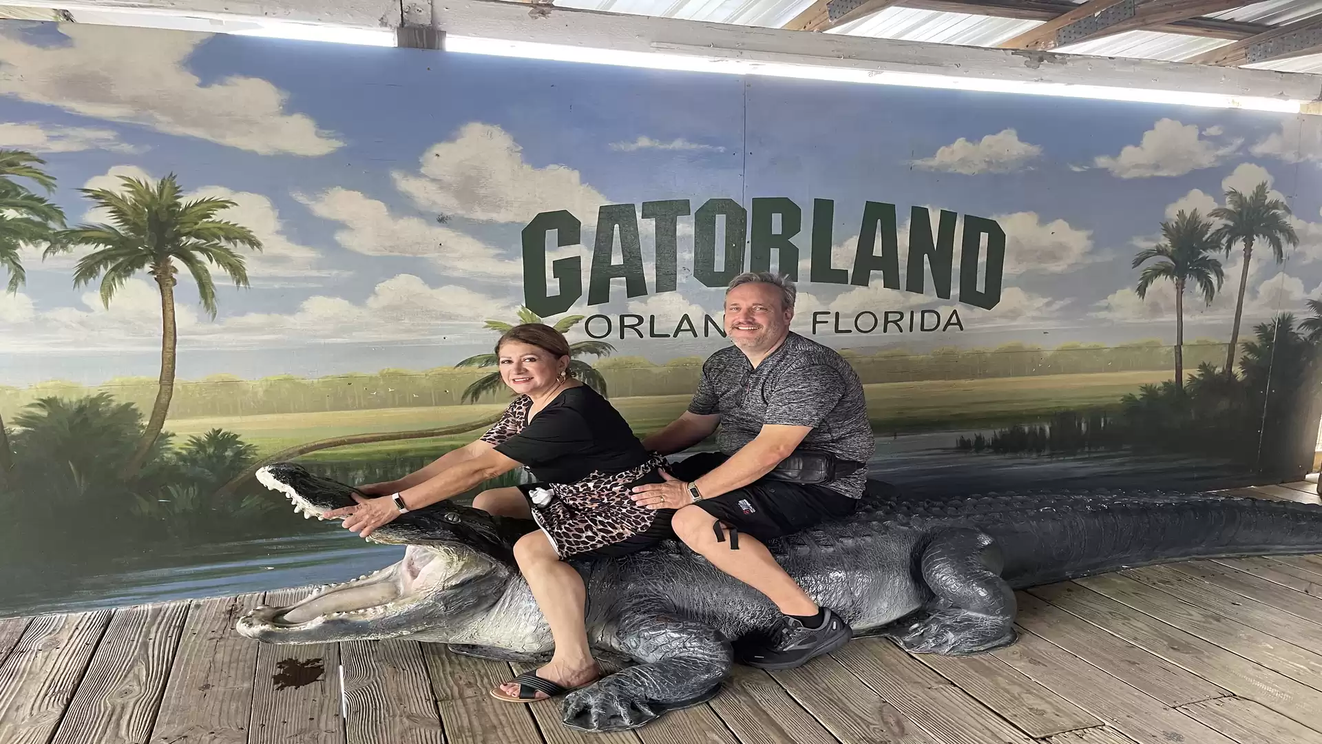 A man and a woman are sitting and laughing on an alligator sculpture in front of a mural with the inscription 'Gatorland Orlando Florida'.