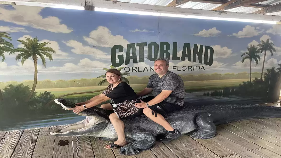 A man and a woman are sitting and laughing on an alligator sculpture in front of a mural with the inscription 'Gatorland Orlando Florida'.