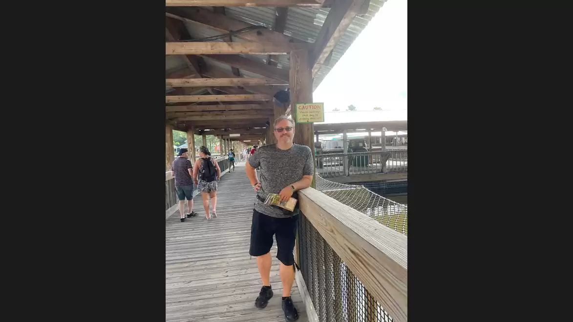 A man stands relaxed on a wooden walkway under a covered area. Other people are visible in the background.