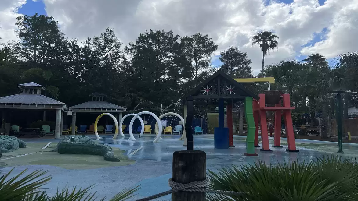 A water park with various colorful play structures and water jets, surrounded by trees and a cloudy sky.