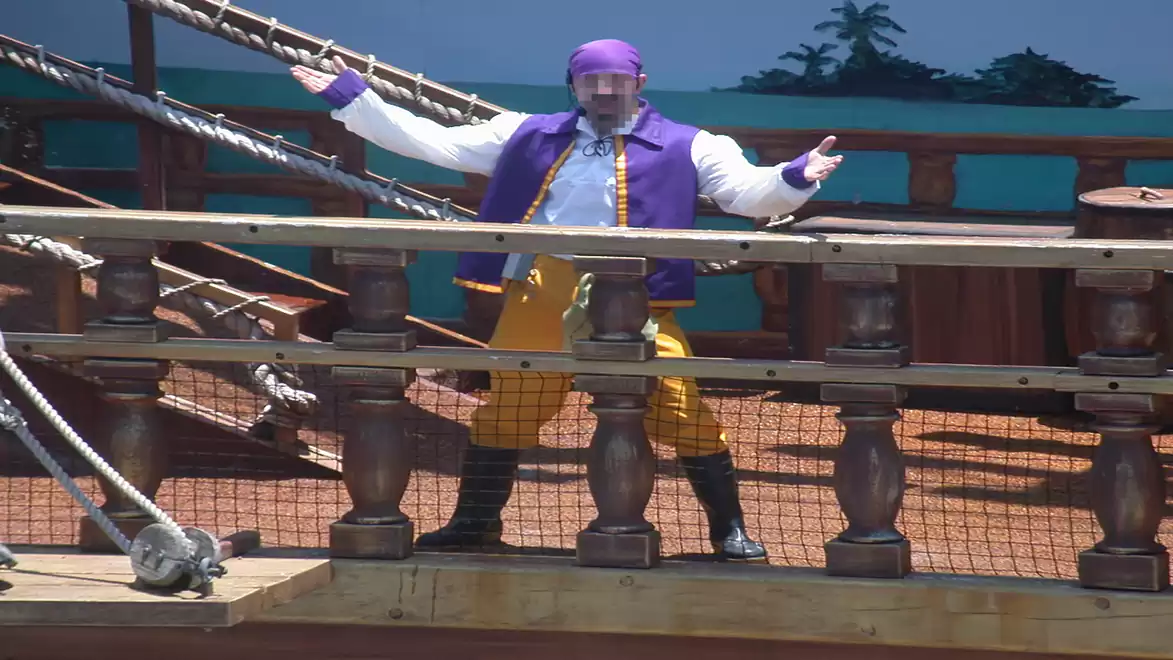 A man in yellow pants and purple vest stands with open arms on a wooden ship deck.