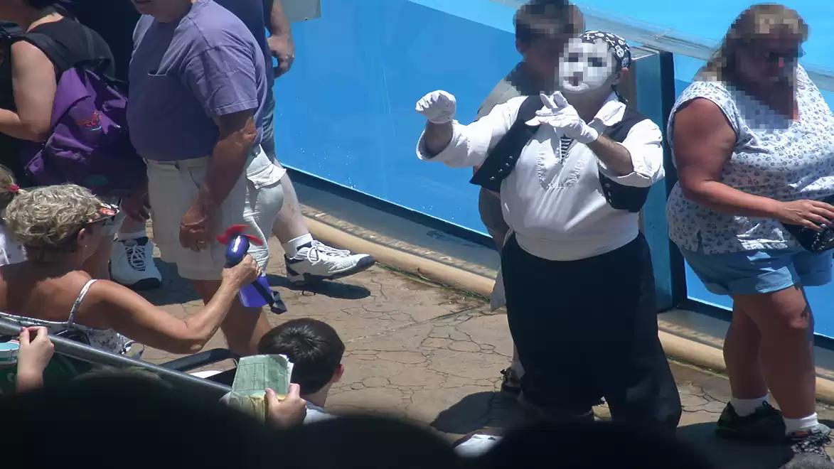 A mime artist in white clothing and gloves entertains an outdoor crowd. Some spectators take photos and videos while others pass by.