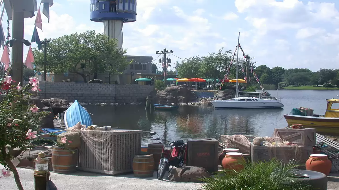 A picturesque harbor on a sunny day with colorful boats and a lighthouse in the background. Nets and pots are visible in the foreground.