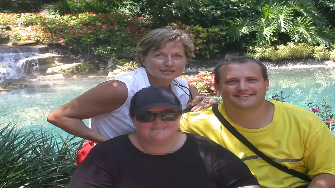 Three people pose outdoors in front of a tranquil pond with lush vegetation and a small waterfall under the sunlight.