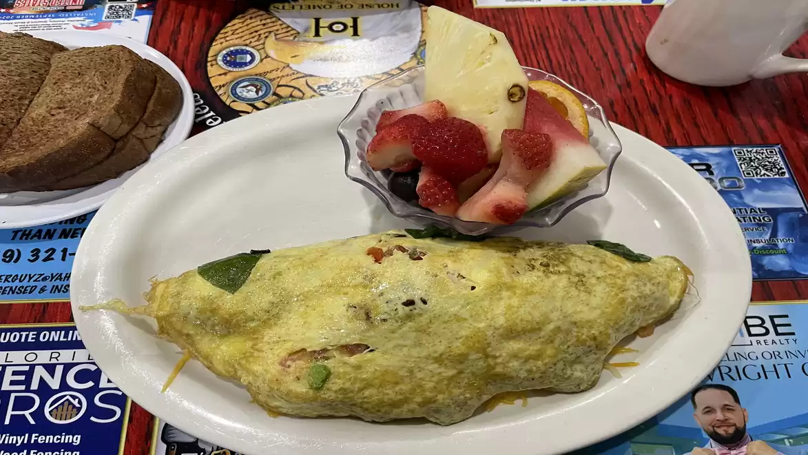 An omelet on a plate with a bowl of fresh fruit including melons, strawberries, and grapes, next to a plate with bread.