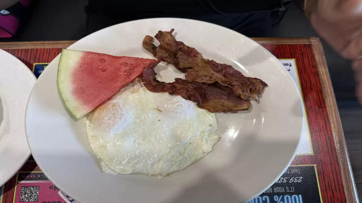 A breakfast plate with two fried eggs, two strips of crispy bacon, and a slice of watermelon on a white plate.