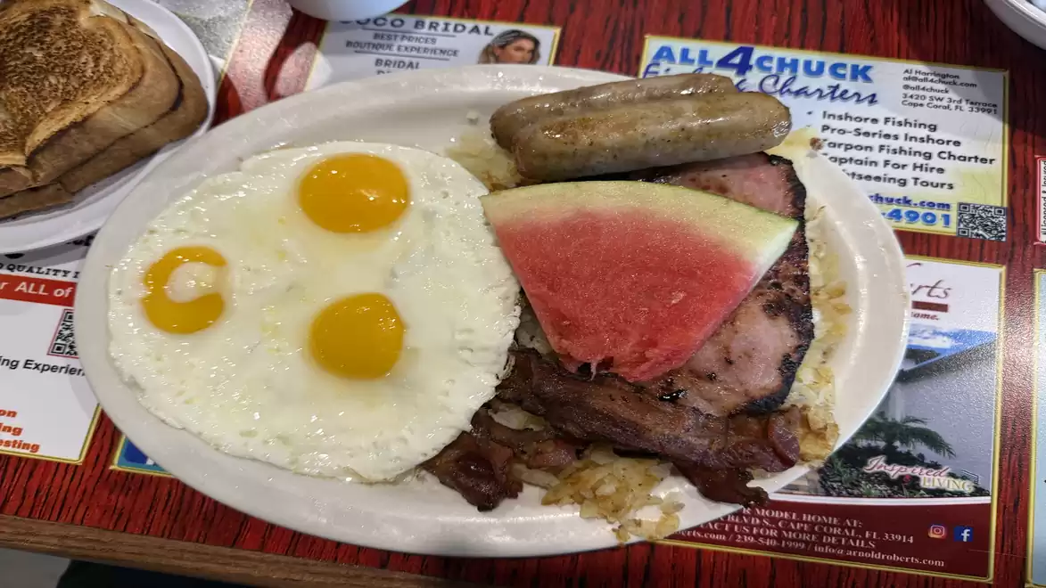 A breakfast plate with fried eggs, sausage, ham, and a slice of watermelon.