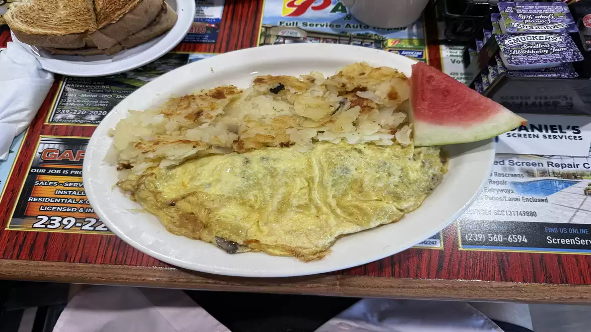 A plate with an omelet, hash browns, and a slice of watermelon on a table in a restaurant.