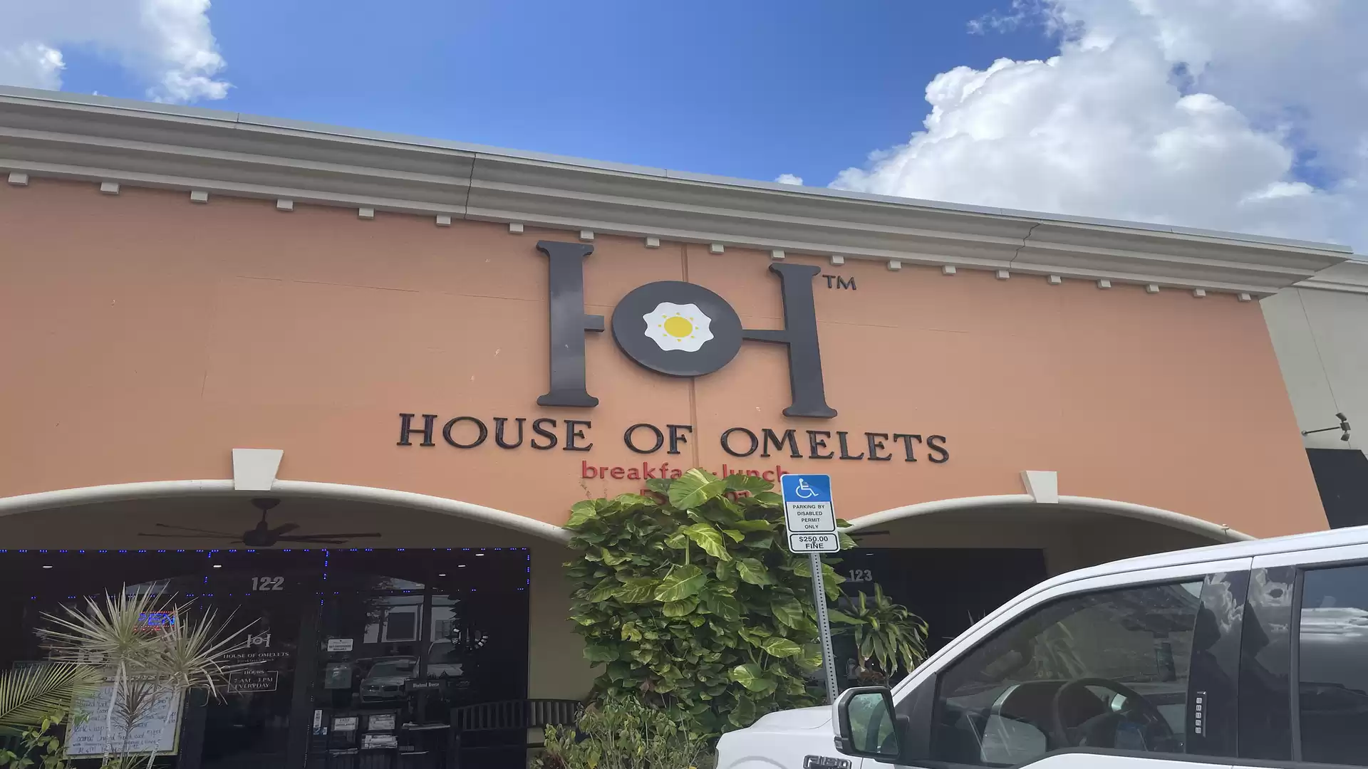 Exterior view of a building with the sign 'House of Omelets'. A car is visible in the foreground, and the sky is blue with a few clouds.