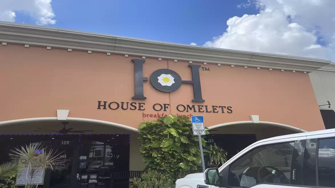Exterior view of a building with the sign 'House of Omelets'. A car is visible in the foreground, and the sky is blue with a few clouds.