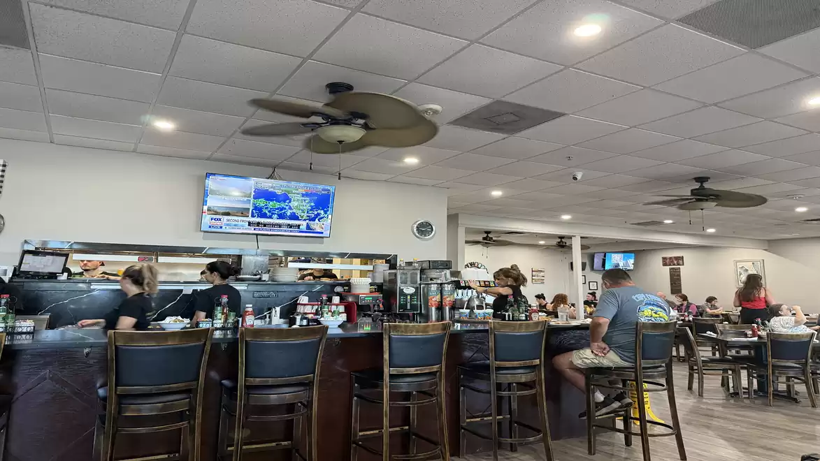 A cozy café with a bar counter where several people are seated. In the background, a TV, ceiling fans, and various seating areas are visible.