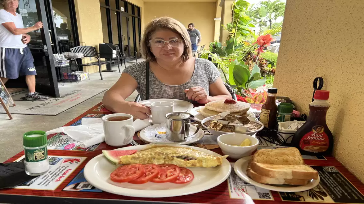 A woman sits at an outdoor table with breakfast including an omelet, tomatoes, toast, coffee, and syrup. A restaurant with flowers and plants is visible in the background.
