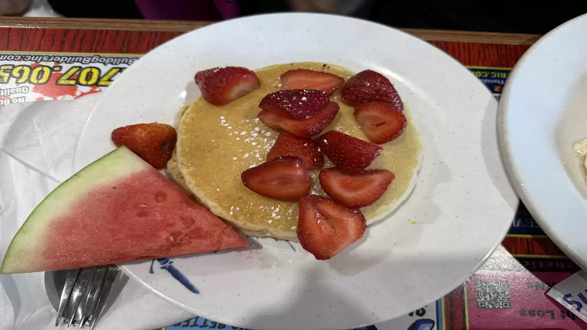 A plate with a pancake topped with sliced strawberries and honey, next to a large piece of watermelon.