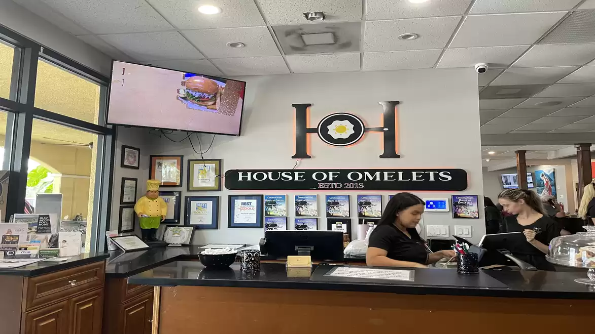 Interior view of a restaurant named 'House of Omelets'. A counter with staff, wall decorations, and a screen displaying a dish.