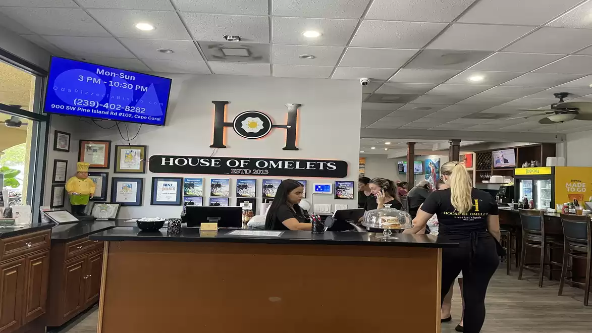 Interior view of a restaurant with several staff members behind a reception counter. A sign with the restaurant's name is mounted on the wall. Guests can be seen in the seating area in the background.