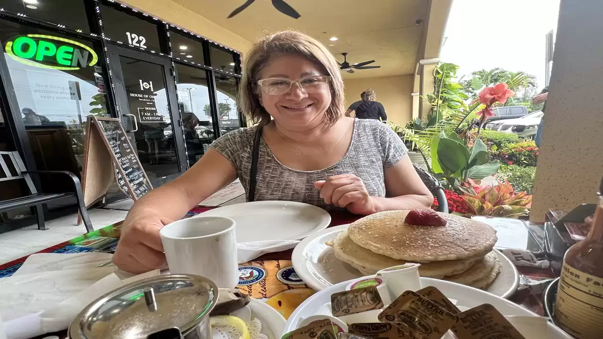 A woman sits smiling in front of a café, ready to enjoy a breakfast with pancakes.