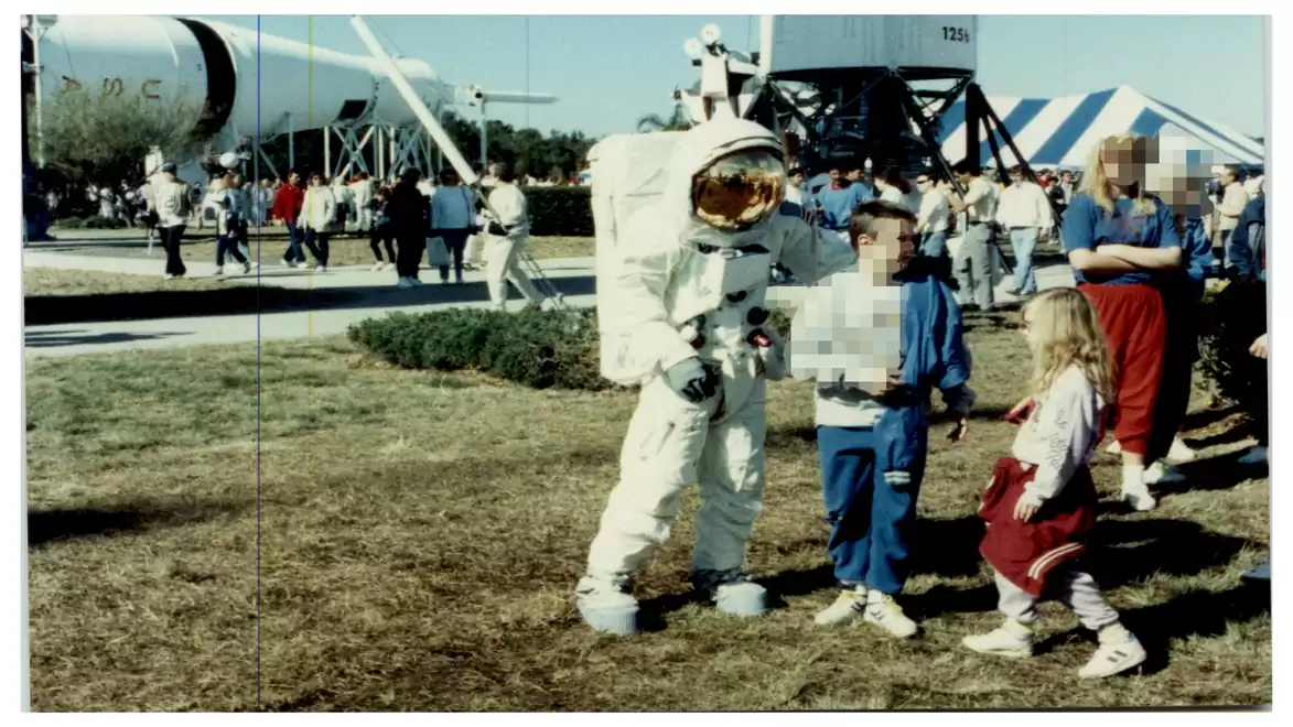 An astronaut in a spacesuit poses with a boy and a girl in an outdoor museum filled with spacecraft and rockets.