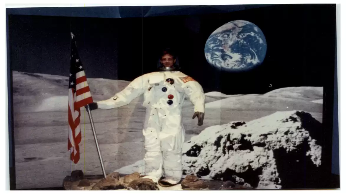 A person in a spacesuit standing on a lunar surface next to an American flag. The Earth is visible in the background.