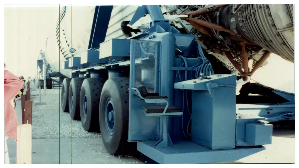A large blue transport vehicle carries a heavy, cylindrical load reinforced with metallic structures.