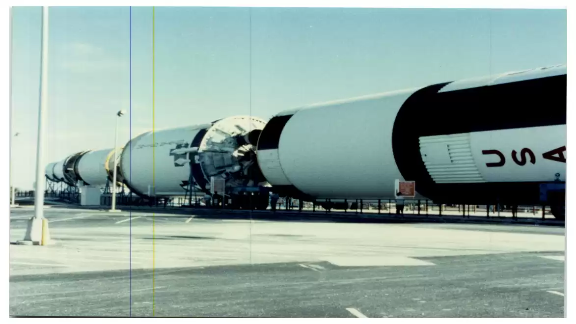 A large, horizontally laid rocket body, apparently from a space model, can be seen on an open site. The rocket's surface is white with black markings and the lettering 'USA' is visible on it.