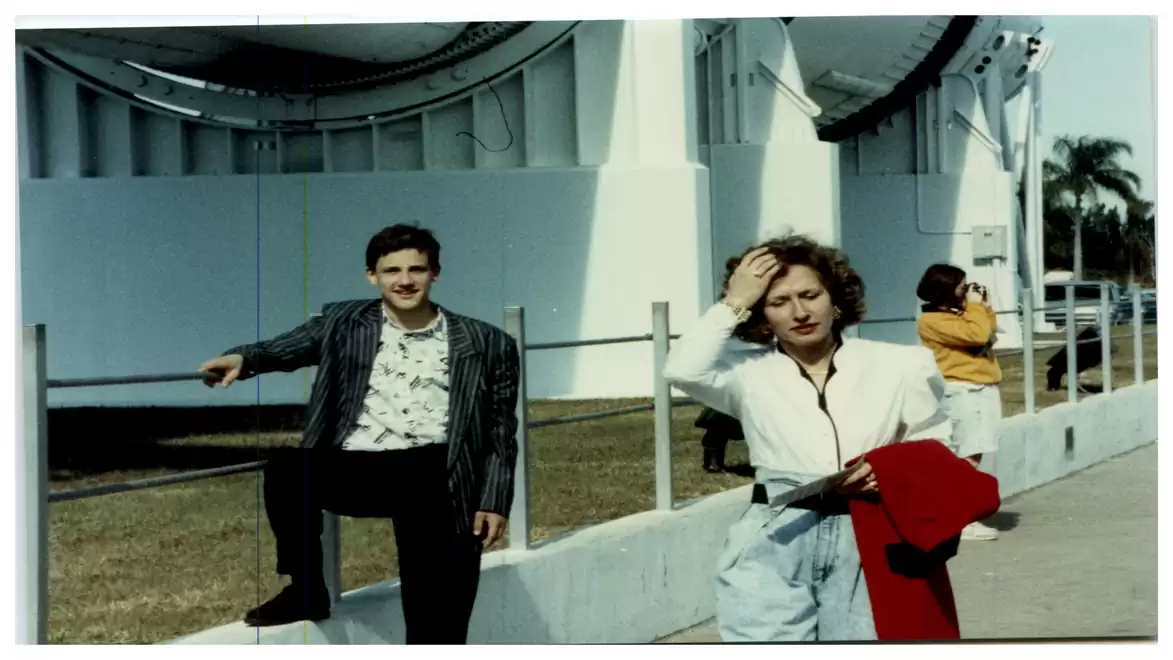 A man in a striped blazer leans casually on a barrier. A woman with a red jacket in hand stands next to him, holding her hand to her forehead. In the background, there is a building and a person in yellow clothing.