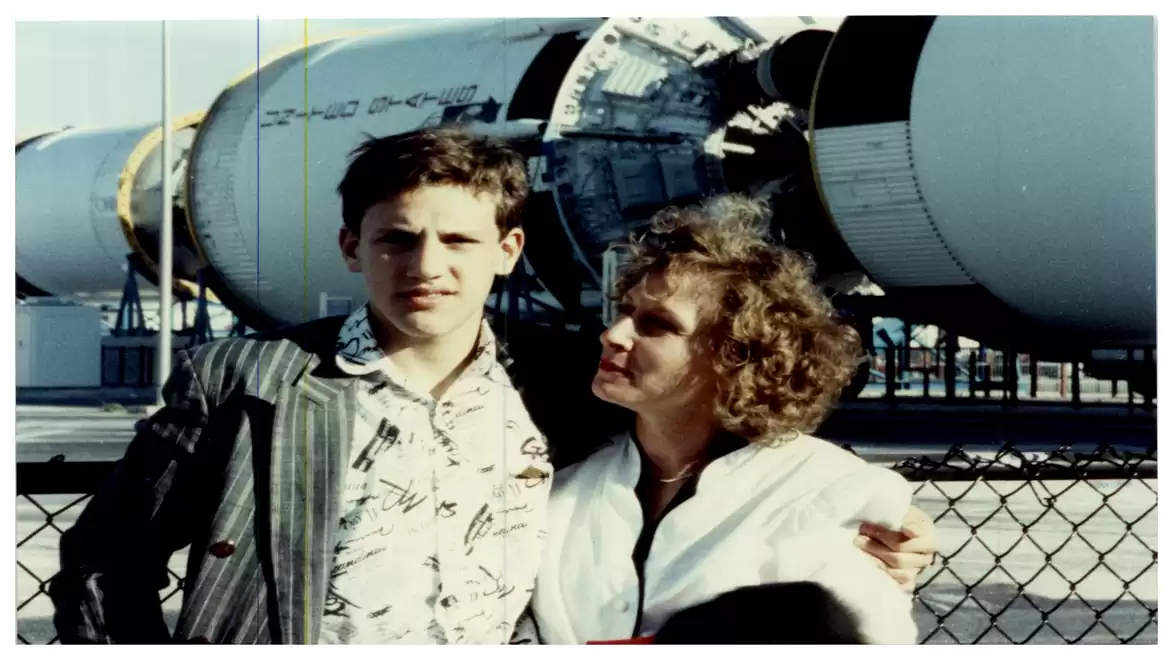 A man and a woman are standing in front of a large rocket in an outdoor setting. Both are casually dressed and look at the camera.