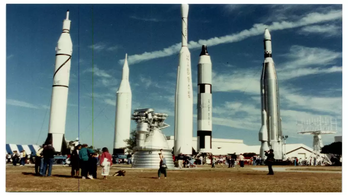 An outdoor exhibit of rockets with several vertically positioned space rockets and visitors on a sunny field.