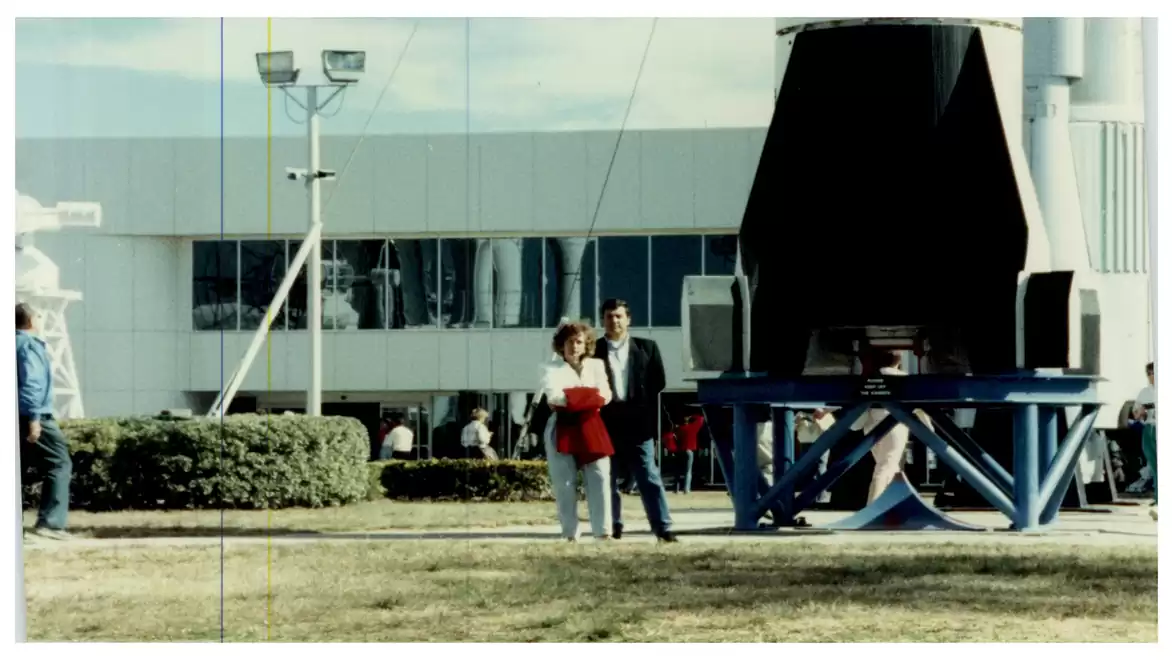 Two people standing outside in front of a technical building with a large machine structure in the background.