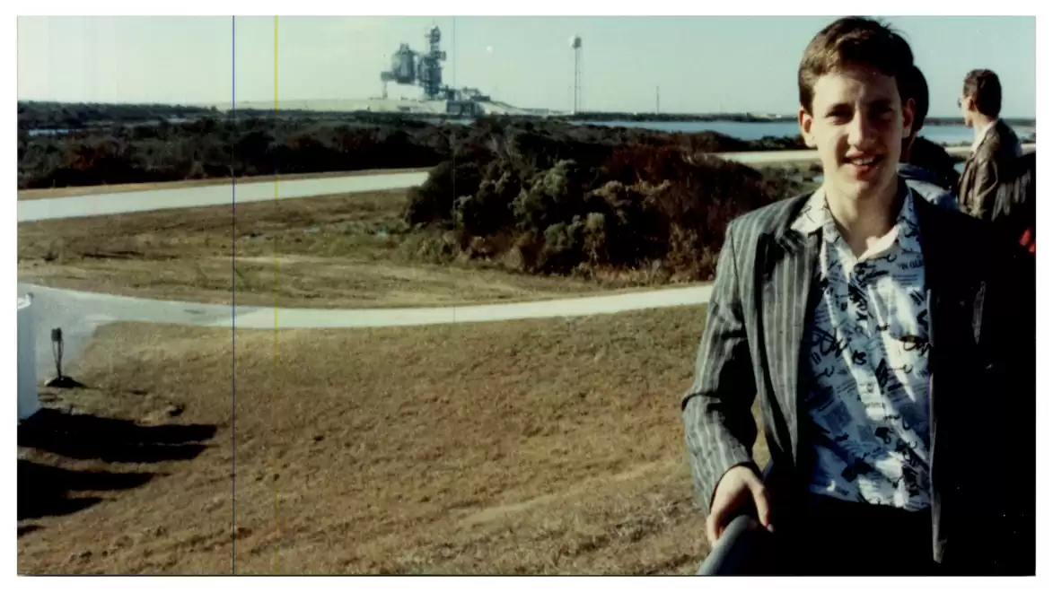 A young man stands smiling in the foreground of a rural landscape with a large industrial structure in the background.