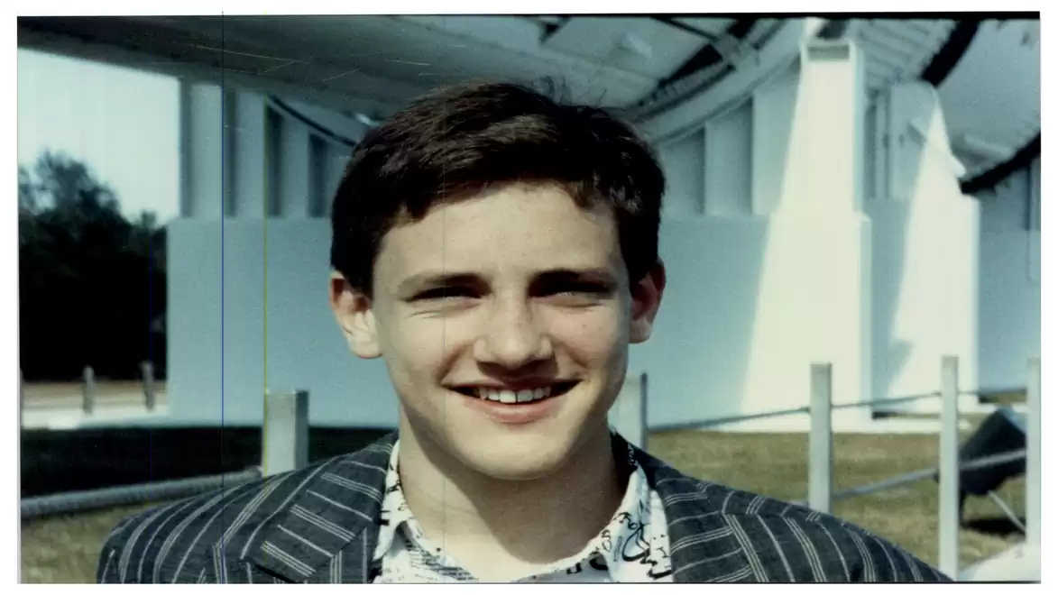 A young man is standing outdoors, smiling in front of a large building or structure. He is wearing a striped blazer and a patterned shirt.