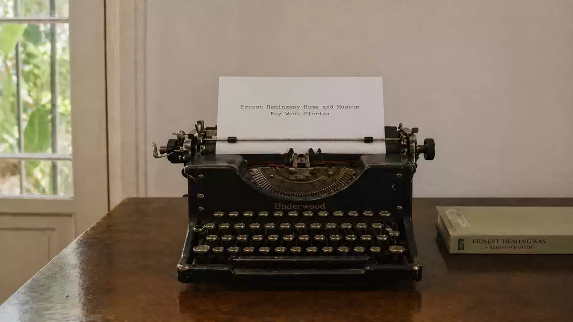 Old typewriter on a wooden table with a sheet of paper and a book next to it. Window in the background shows trees.
