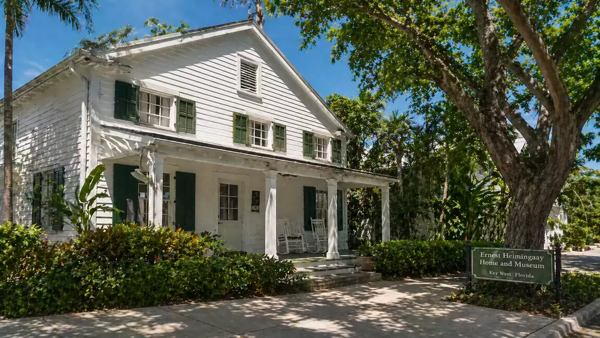 The image shows a white historic wooden house with green shutters, surrounded by lush trees. In front of the house, there is a wooden sign with information.