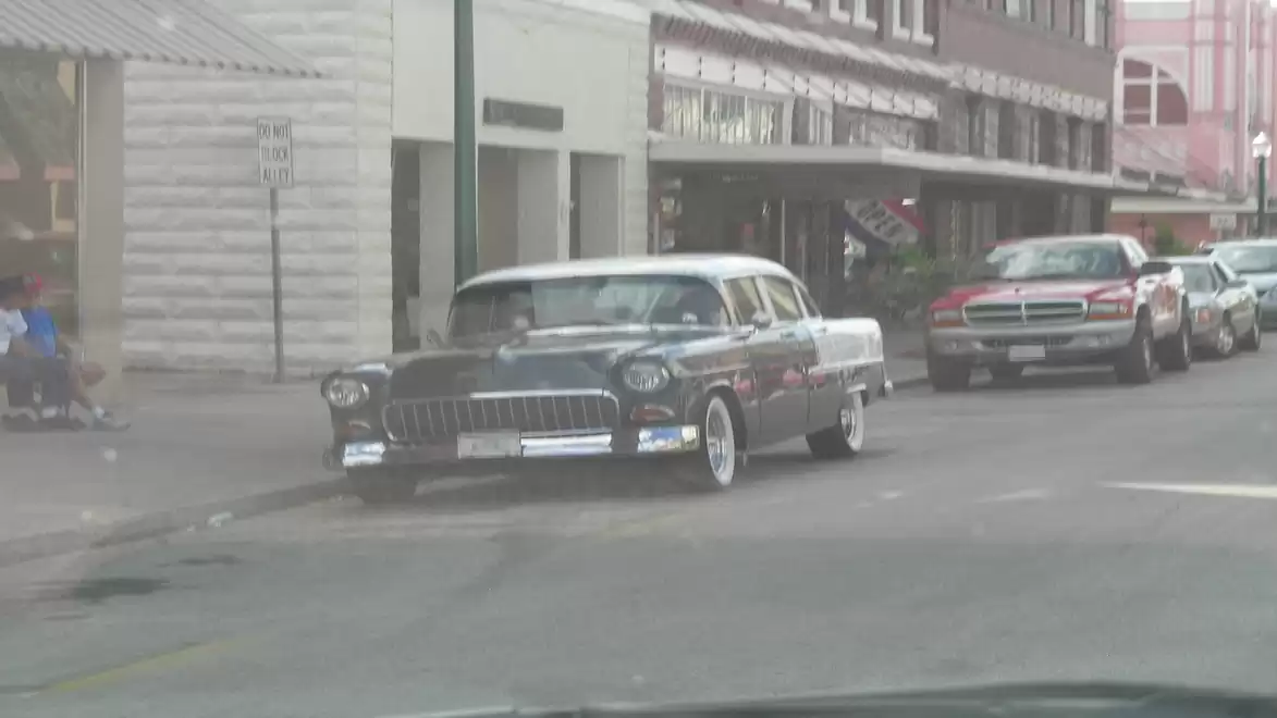 A classic car parked on a street in an urban setting with several buildings in the background.