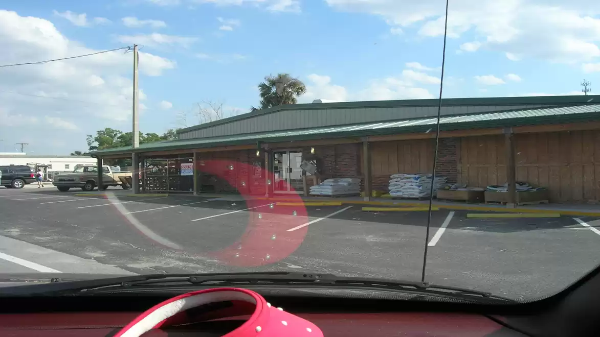 An empty parking lot in front of a store with a green roof; in the foreground a red dashboard with a polka dot bag.