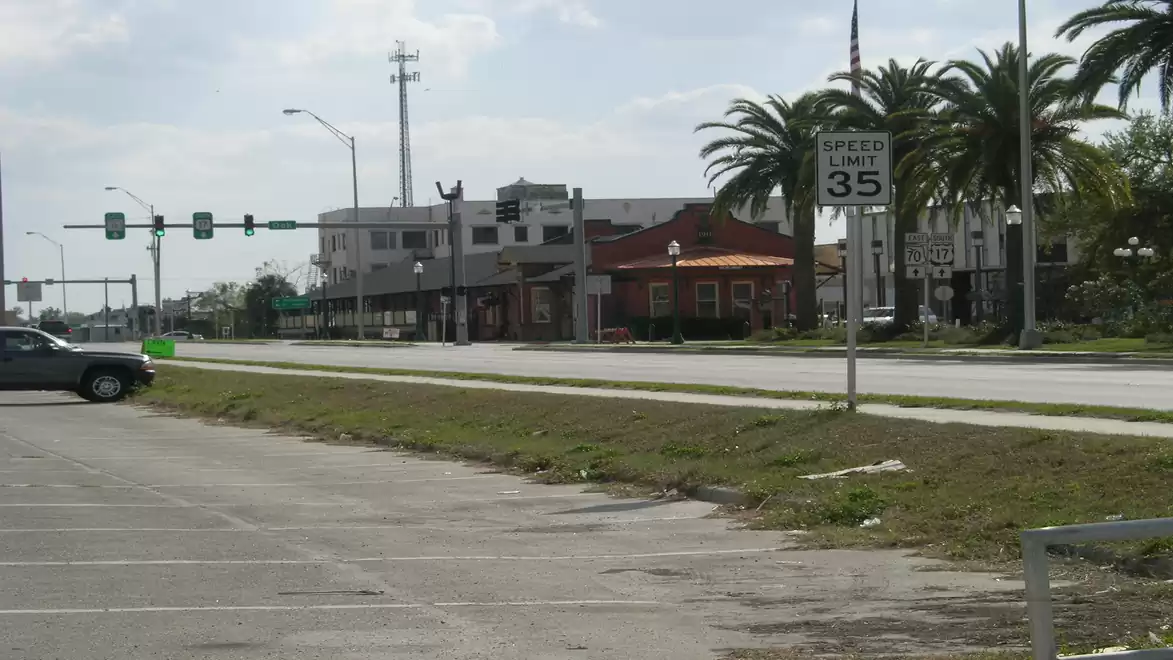 Empty urban street with speed limit sign, scattered palm trees, and buildings in the background on a sunny day.