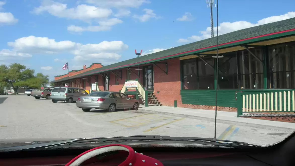 View of a parking lot with several cars in front of a long brick building with a green roof and blue sky in the background.