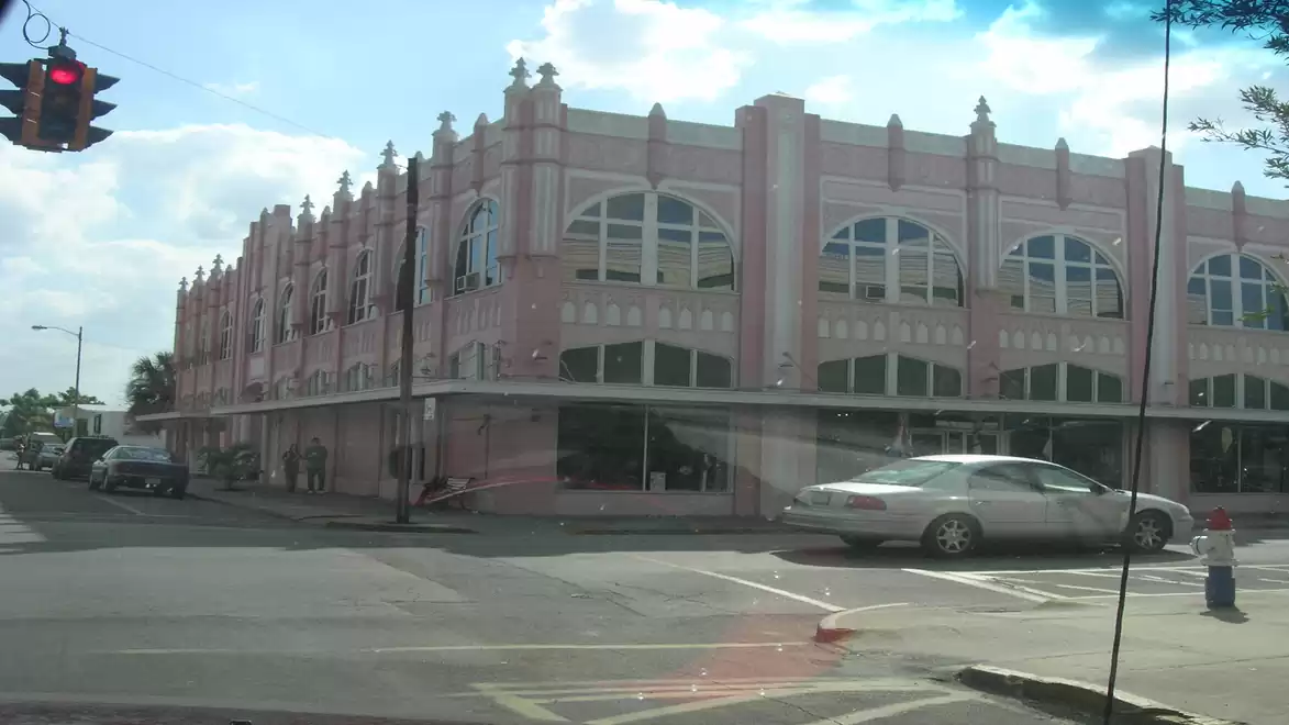 A pink Art Deco style building at a street corner with cars in the foreground and a cloudy sky in the background.