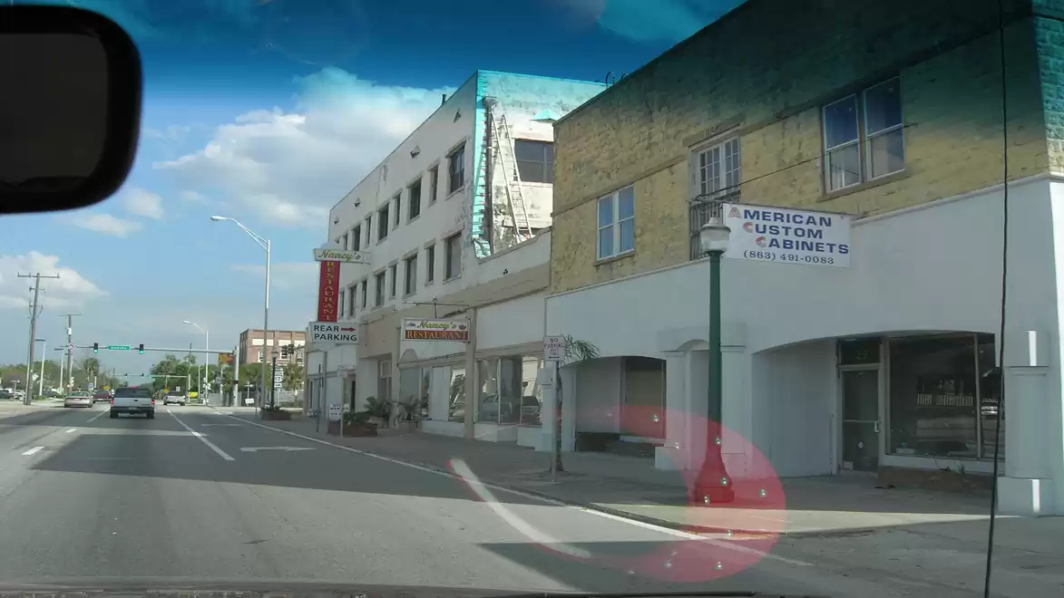 View from a car windshield onto a quiet town street with multi-story buildings and a sign indicating American cabinets.
