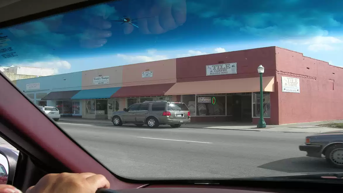 View from a car window showing a street scene with shops and parked cars under a blue sky.