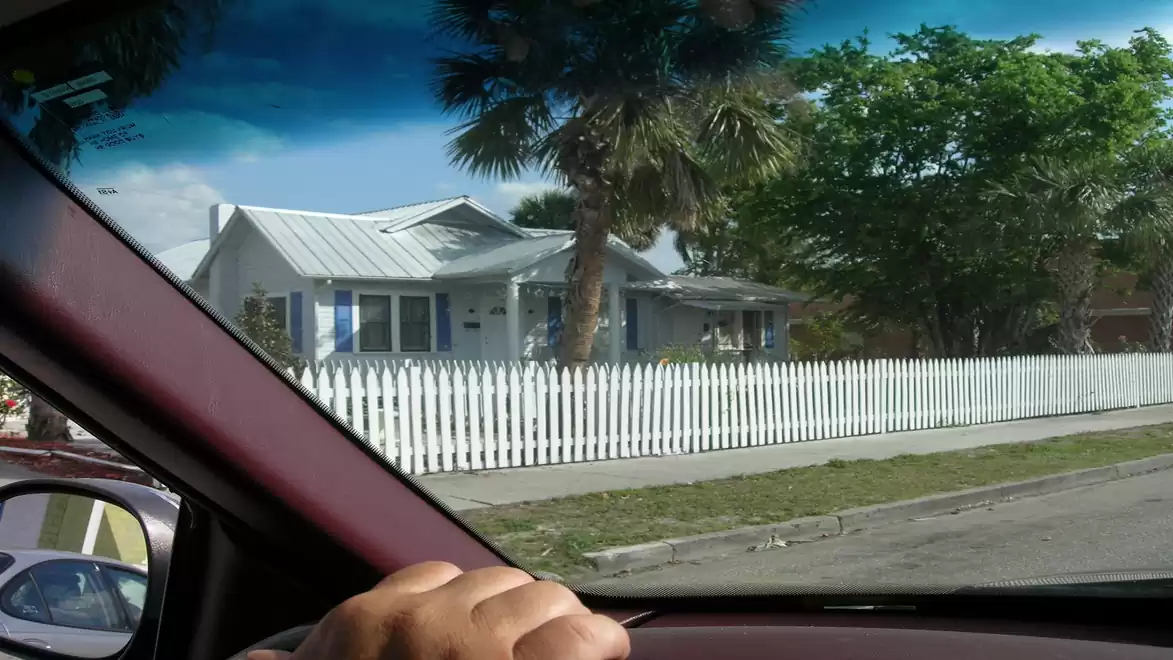 View from a car through the windshield towards a white house with a white picket fence and palm trees in the foreground.