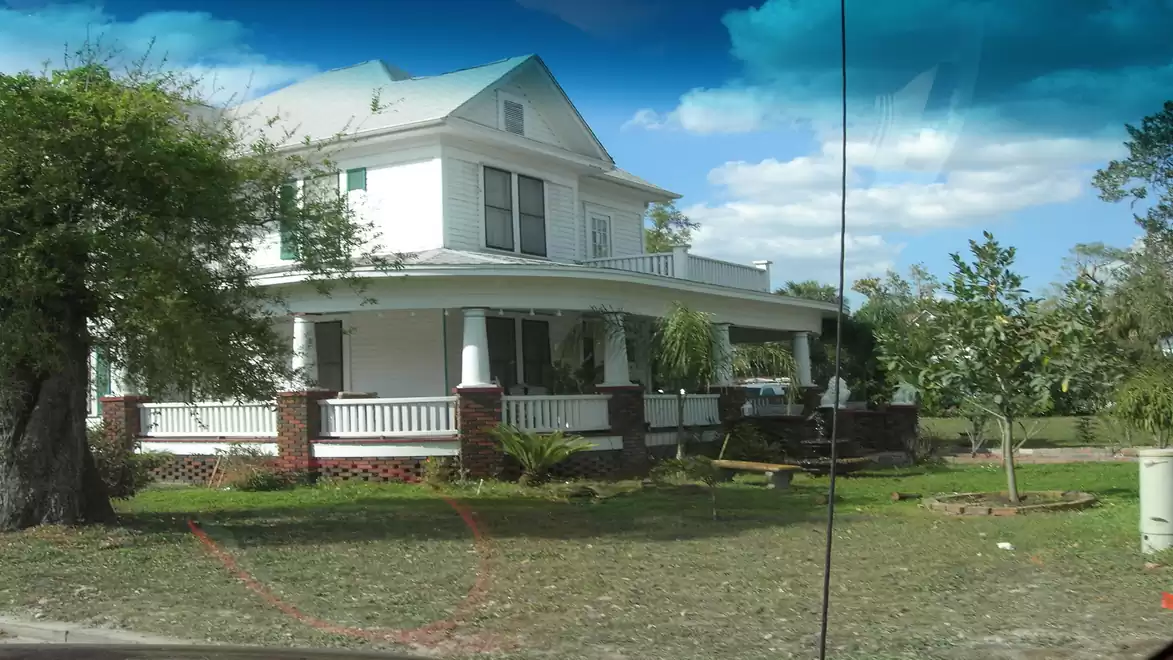 A large white house with a porch surrounded by green lawn and trees under a blue sky.
