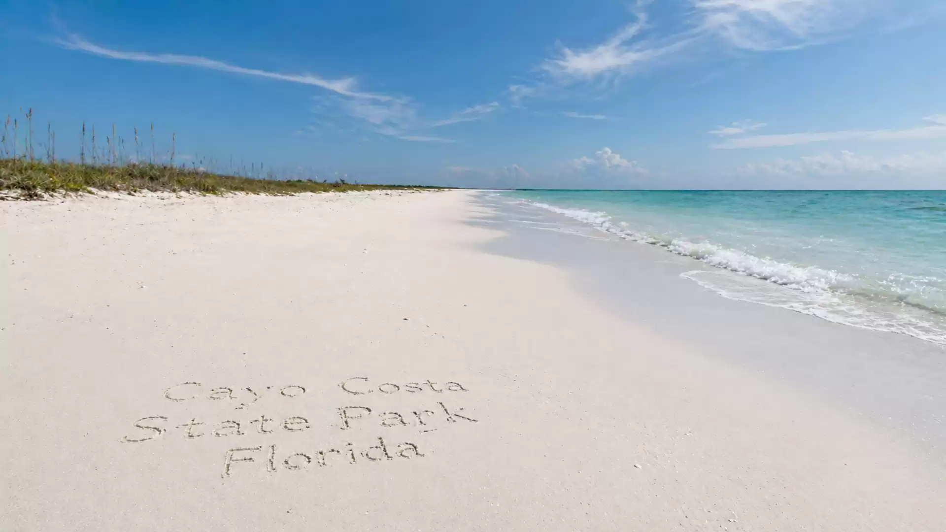 A wide, pristine sandy beach at Cayo Costa State Park, Florida, with clear turquoise waves gently lapping at the shore. The sky is blue with a few clouds.