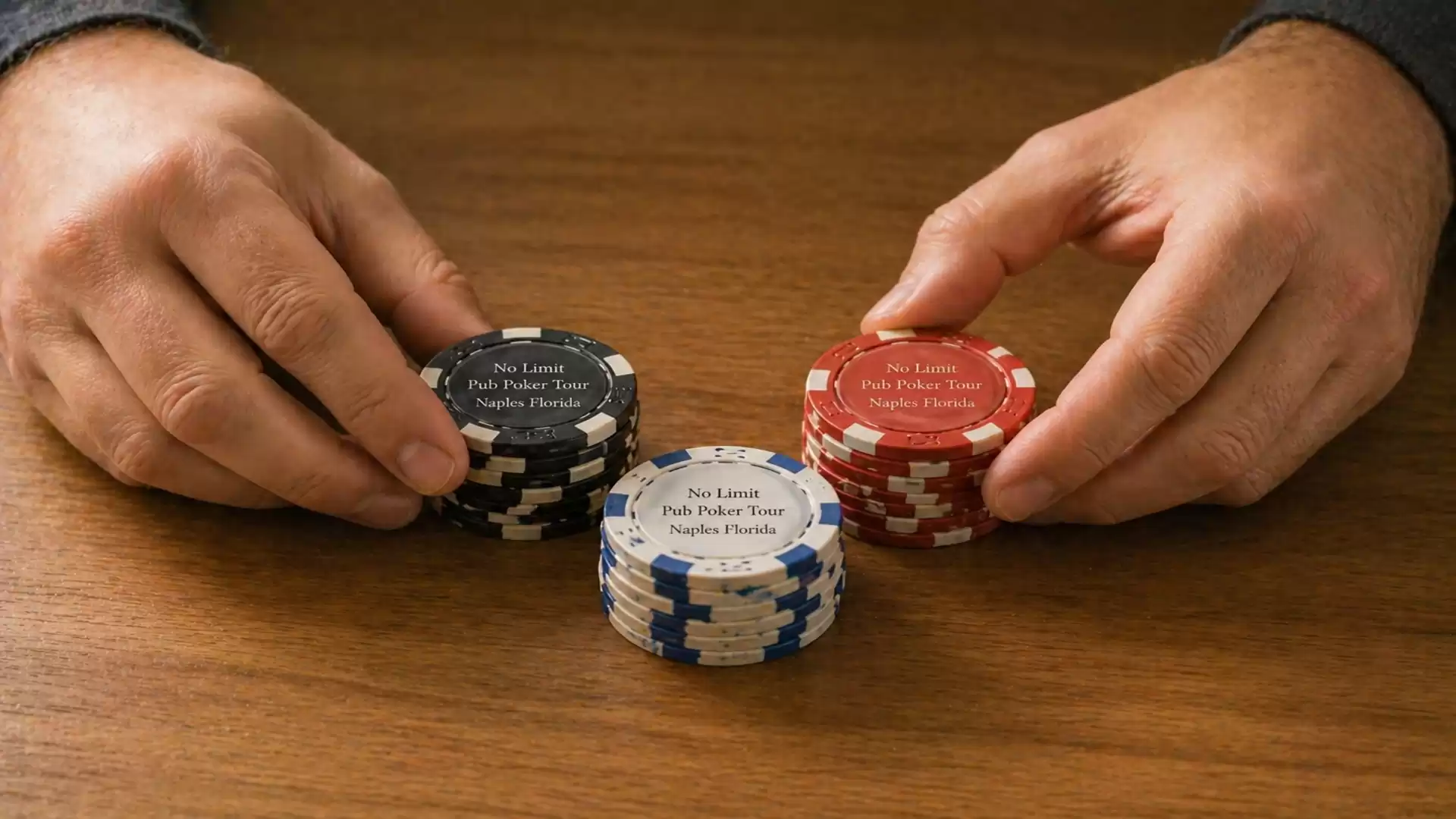 Two hands holding poker chips in different colors - black, blue, red - on a wooden table.