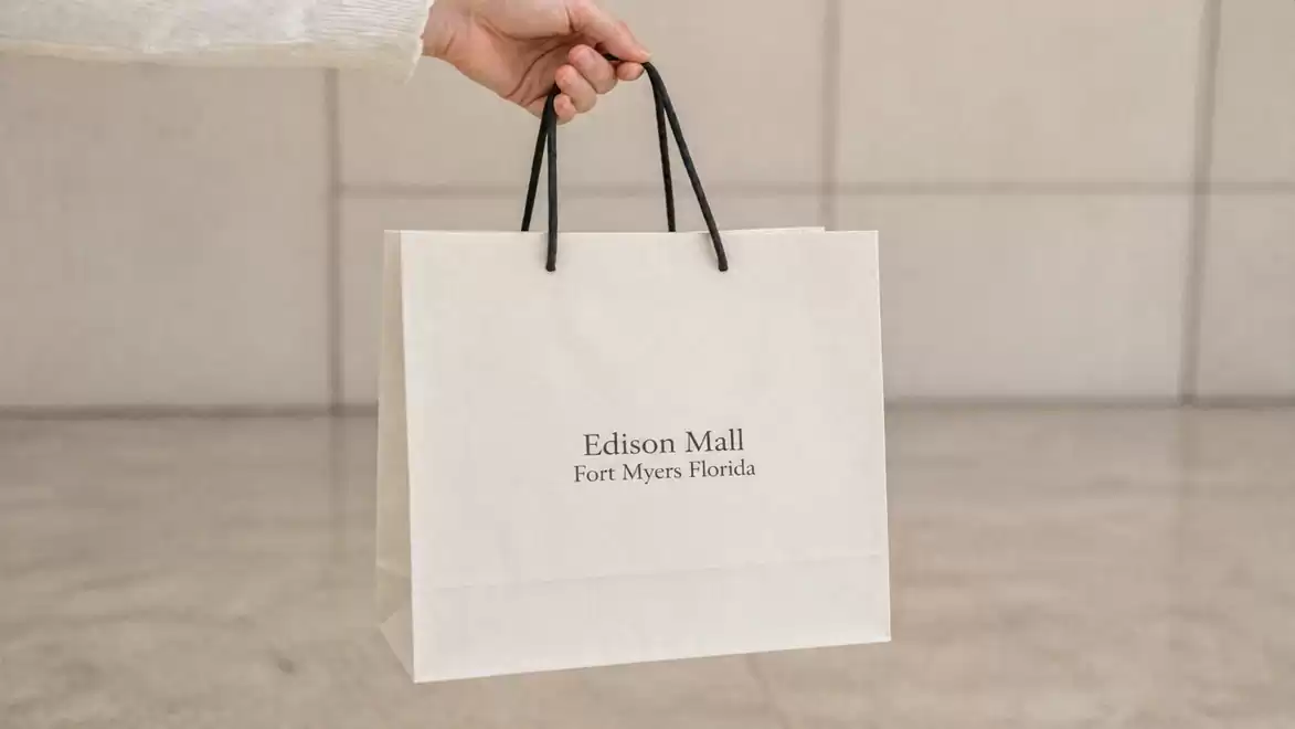 A hand holds a shopping bag with the inscription 'Edison Mall, Fort Myers Florida'. The bag has black handles and is in a minimalist space.
